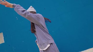 Genç bir kadın enerjik hip hop dansı yapıyor, gri eşofman ve bere giyiyor, canlı mavi duvar arka planına karşı kentsel tarzını ifade ediyor.
