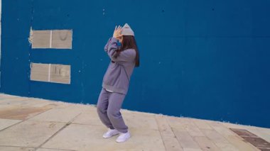 Kadın dansçı dinamik hip hop koreografisi yapıyor, canlı mavi şehir duvarına karşı sanatsal enerjisini ifade ediyor, çağdaş sokak dansı becerisi ve tutkulu hareket sergiliyor.