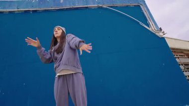 Kendine güvenen kadın dansçı dinamik hip hop koreografisi sergiliyor, canlı mavi duvar arkaplanına karşı kentsel enerji ve yaratıcılığını ifade ediyor