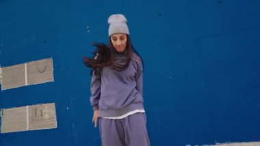 Dinamik kadın dansçı, dinamik hip hop koreografisini icra ediyor, canlı mavi duvar arka planına karşı ritmik hareketler ve şehir tarzı sergiliyor.