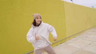 Dinamik kadın dansçı, parlak sarı duvara karşı yüksek enerjiyle hip hop koreografisi yapıyor, akışkan hareketler ve şehir ritmi yoluyla sokak dansı tarzını ifade ediyor.