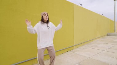 Dinamik kadın dansçı, sarı şehir duvarına karşı canlı enerjiyle hip hop koreografisini icra ediyor, çağdaş sokak dansı hareketini ve kişisel sanatsal ifadesini somutlaştırıyor.