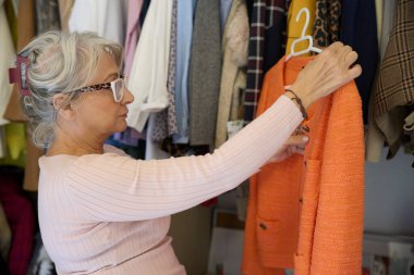 Son sınıf öğrencisi şık bir kadın. Kıyafet dolabından turuncu bir ceket seçiyor. Moda seçimi yapıyor.