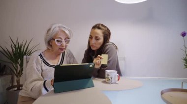 Olgun bir kadın, destekleyici kızıyla online alışveriş yapmayı öğreniyor, sıcak bir bağlantı anını paylaşırken dijital teknolojiyi birlikte kullanıyor.