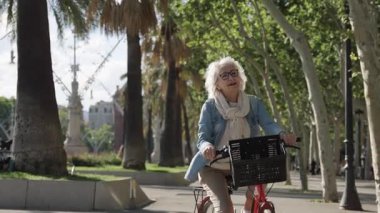 Yaşlı bir kadın parkta bisiklet sürüyor, güneşli bir öğleden sonra neşe ve özgürlükle pedal çeviriyor, sakin kentsel keşiflerle aktif bir kıdemli yaşam tarzı benimsiyor.