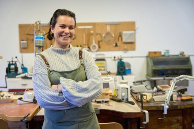 Atölyesinde çapraz kollarla poz veren gülümseyen bir mücevher sanatçısı portresi sanatında güven ve uzmanlık gösteriyor.