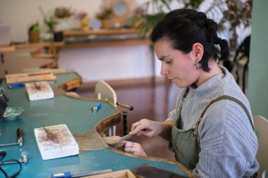 Kadın kuyumcu, atölyesindeki bir metal parçasını dikkatlice düzelterek, el yapımı mücevherler yaratıyor.