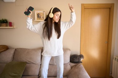 Kulaklıkları takılı ve akıllı telefonları elinde, genç bir kadın kanepesinde neşeyle dans ediyor, oturma odasında müziğin keyfini çıkarıyor.