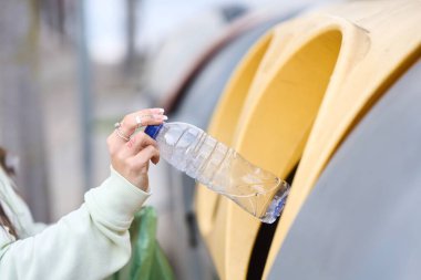 Sarı geri dönüşüm kutusuna plastik şişe atan bir kadın, çevresel sorumluluk ve atık ayıklamayı teşvik ediyor.