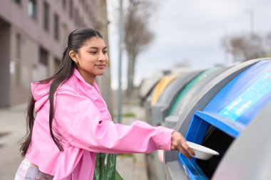 Plastik kaseyi geri dönüşüm kutusuna atan genç bir kadın, çevre bilincini ve sorumlu atık yönetimini teşvik ediyor.
