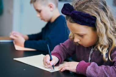 İlkokul öğrencileri canlı bir sınıf ortamında derslerine yoğunlaşırken not defterlerine yazmakla meşguller.
