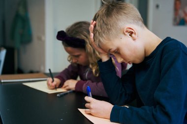 Erkek ve kız kardeş bir masada oturup ödevlerini yapıyorlar, çocuk yorgun görünüyor ve başını elinin üzerine koyuyor.
