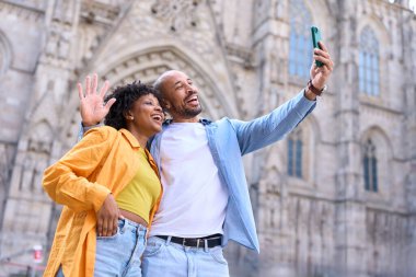 Neşeli çift yaz tatili boyunca çarpıcı Barselona Katedrali 'ne el sallarken selfie çekiyor, canlı şehrin tadını çıkarıyorlar.