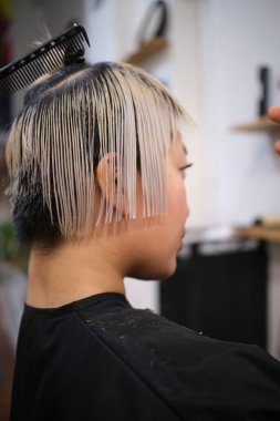 Kuaför, bir kuaförde kısa sarı saçlı genç bir kadının ıslak saçlarını tarıyor ve kesiyor.