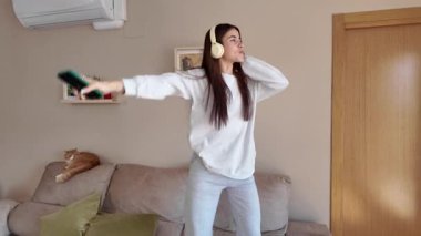 Kablosuz kulaklık takan bir kadın oturma odasındaki kanepede enerjik bir şekilde dans ediyor, akıllı telefondan kaygısız, neşeli hareketlerle müziğin tadını çıkarıyor.