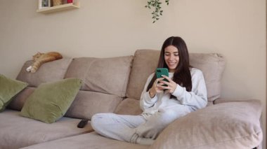 Evdeki rahat koltukta otururken akıllı telefon kullanan rahat genç kadın, arka planda huzur içinde uyuyan kızıl kedi.