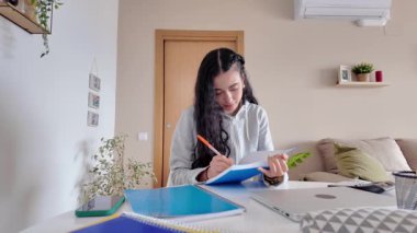 Öğrenci yaklaşan sınavlara hazırlanırken özenle notlar yazıyor, odaklanmış konsantrasyonla rahat bir evde özenle masa başında çalışıyor.