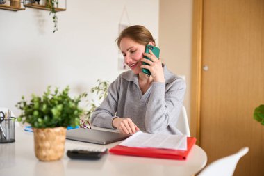 Gülümseyen serbest çalışan kadın işini evden yönetiyor, telefonla konuşuyor ve masasındaki belgeleri kontrol ediyor.