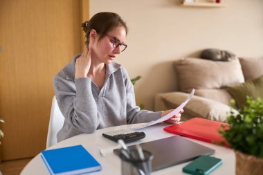 Muhasebeci kadın evrakları okuyor ve ev işleriyle uğraşırken boynuna masaj yapıyor. Son teslim tarihlerinin verdiği stresi hissediyor.