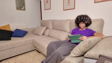 Kanepede dinlenen genç öğrenci çevrimiçi öğrenme, eğlence ve iletişim için dijital tablet kullanıyor.