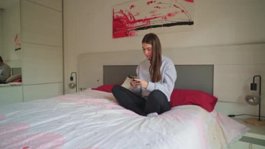Yatakta rahat pijamalı genç bir kadın rahat yatak odasının içinde akıllı telefonlara bakıyor.