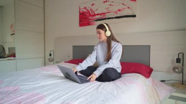 Konforlu yatak odasından uzaktan çalışan, kulaklık takan, bilgisayar ve akıllı telefonu online öğrenme ve serbest çalışma görevleri için kullanan genç bir kadın.