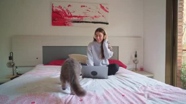 Uzaktan çalışırken dizüstü bilgisayara odaklanan genç bir kadın, yatak odasının yanında yürüyen evcil bir kedi, modern dijital yaşam tarzı ve rahat bir ev ortamı sunuyor.
