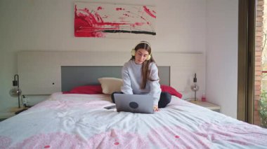 Kulaklık takan, internetten öğrenmek için laptop kullanan, rahat bir şekilde yatağında oturan bir öğrenci.