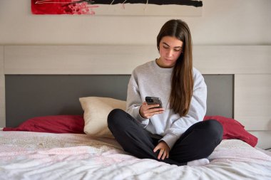 Yatakta bacak bacak üstüne atmış bir kız öğrenci yatak odasında cep telefonuyla sosyal medyada geziniyor.
