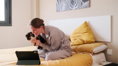 Genç bir kadın yatakta oturuyor, tabletle video görüşmesi yapıyor ve kedisini diğer katılımcılara gösteriyor.