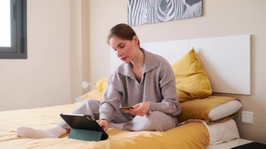 Genç bir kadın online alışveriş için dijital tablet ve kredi kartı kullanarak rahatça yatakta oturuyor.