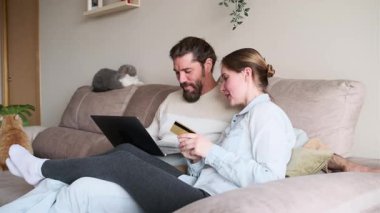 Genç çift kredi kartı ve dizüstü bilgisayarla online alışveriş yaparken gülüyor, kanepede birlikte vakit geçiriyorlar.