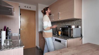 Neşeli erkek, parlak çağdaş mutfakta sağlıklı bir kahvaltı hazırlıyor. Taze malzemeler ve içeceklerle sabah rutininin tadını çıkarıyor.