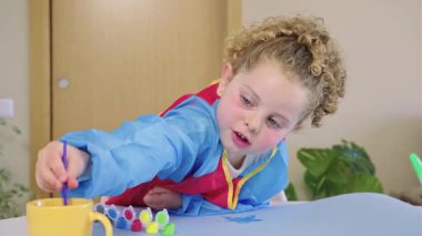 Renkli önlük giyen genç bir kız resim fırçasını dikkatlice suya batırırken evde sanat eserleri yaratıyor ve çocukluk yaratıcılığını ve sanatsal becerilerini gösteriyor.