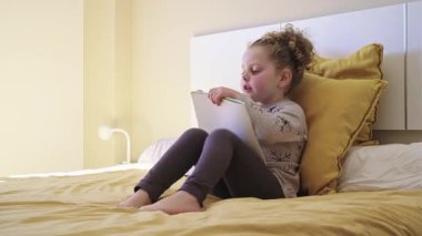Genç kız rahat bir şekilde yumuşak yatakta otururken kitap okuyor. Altın saat ışığında huzurlu bir şekilde öğrenme ve yaratıcı hikaye anlatımının tadını çıkarıyor.