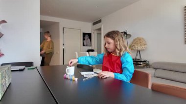 Annesi arka planda bir şeyler hazırlarken, yemek masasında resim yapmaya konsantre olan bir kız.