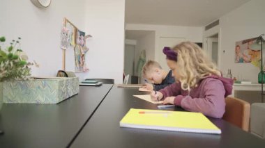 Erkek ve kız kardeş evde birlikte çalışıyorlar, masada oturuyorlar ve defterlerine yazıyorlar.
