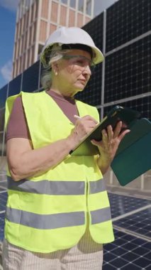 Güneş panellerini yavaş çekimde incelerken tablet ve stil kalem kullanan güvenlik yeleği ve kask giyen kıdemli kadın mühendis.