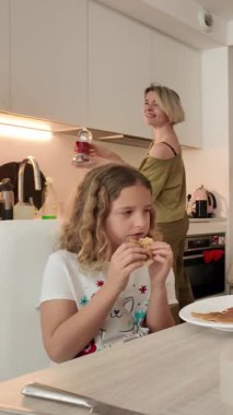 Anne kahve hazırlarken, genç kız aydınlık, modern beyaz bir mutfakta sabah kahvaltısının tadını çıkarırken aile birlikteliğini ve günlük rutinini ön plana çıkarıyor.