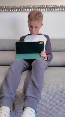 Gri koltukta oturan genç çocuk, evde boş zamanlarında dijital tablet ekranla meşgul olurken şaşkınlık gösteriyor.