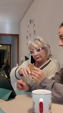 Kızlarıyla online alışveriş yapmayı öğrenen yaşlı bir kadın kredi kartı ve tablet kullanarak yardım ediyor