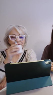 Olgun bir kadın sıcak bir içecek yudumlarken tableti karıştırıyor, sakin bir ifadeyle evde dinleniyor ve doğal gün ışığı yaşam alanından geçiyor.