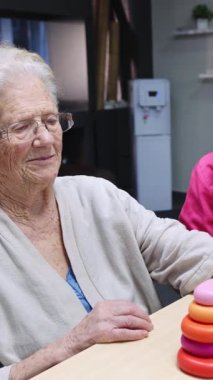 Yaşlı bir kadının, bir hemşirenin yardımıyla yüzükleri istiflediği, neşeli bir rehabilitasyon ve bakım anının gösterildiği yavaş çekim videosu.