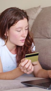 Genç kadın koltukta rahat bir şekilde uzanıyor, kredi kartıyla online alışveriş yapıyor, rahat dijital tüketici yaşam tarzını yavaş çekim perspektifinden yakalıyor.