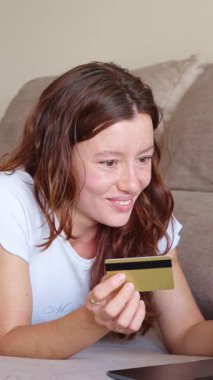 Kanepede uzanan genç bir kadın tablet ve kredi kartıyla online alışveriş yapıyor, ağır çekimde.
