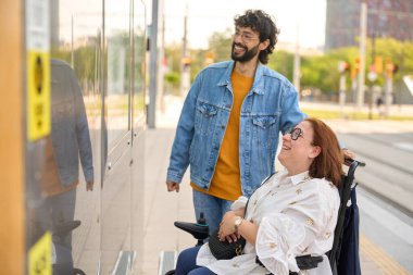 Tekerlekli sandalye kullanan neşeli bir kadına eşlik eden, tramvay durağında bekleyen, paylaşılan mutluluğun ve kentsel ulaşılabilirliğin tadını çıkaran bakıcı.