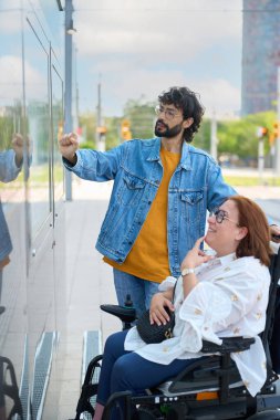 Adam, şehirdeki modern elektrikli sandalyede oturan gülümseyen engelli bir kadınla konuşurken bilgi panelini işaret ediyor.