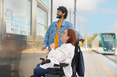 Tramvay ya da tren istasyonunda haritaya bakan turist çift, tekerlekli sandalye kullanan kadın, modern ulaşılabilir seyahat