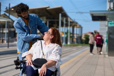 Adam, modern bir tramvay istasyonunda tekerlekli sandalye kullanan gülümseyen bir kadını destekliyor, arkadaşlık ve ulaşılabilirliği somutlaştırıyor.
