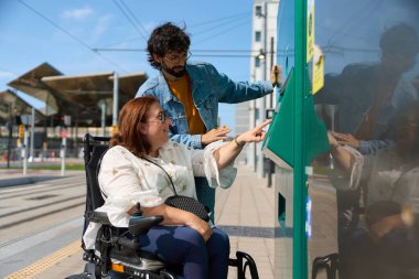 Genç adam tekerlekli sandalyedeki engelli bir kadına bilet alması için yardım ediyor.